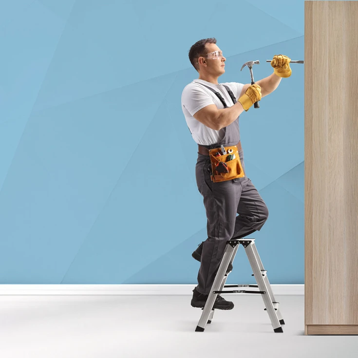 A handyman wearing safety gear and holding a hammer and chisel, working on a wooden surface while standing on a step ladder, symbolising craftsmanship and professionalism, against a blue geometric background.