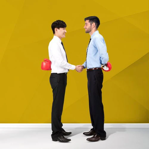 Two men in business attire shake hands while each hides a boxing glove behind their back, symbolising conflict resolution. The background is a bright yellow geometric design, with text on the left reading ‘Dispute Assist’ followed by ‘Resolving disputes, before they get out of hand.’