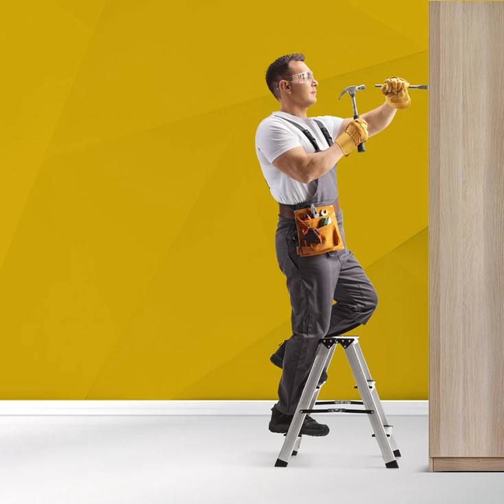 A handyman wearing safety gear and holding a hammer and chisel, working on a wooden surface while standing on a step ladder, symbolising craftsmanship and professionalism, against a blue geometric background.