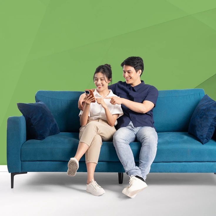 A cheerful couple sitting on a blue sofa, smiling and engaging with a smartphone, symbolising connection and shared experiences, against a blue geometric background.