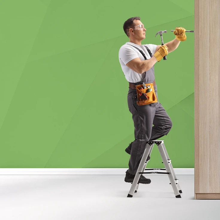 A handyman wearing safety gear and holding a hammer and chisel, working on a wooden surface while standing on a step ladder, symbolising craftsmanship and professionalism, against a blue geometric background.