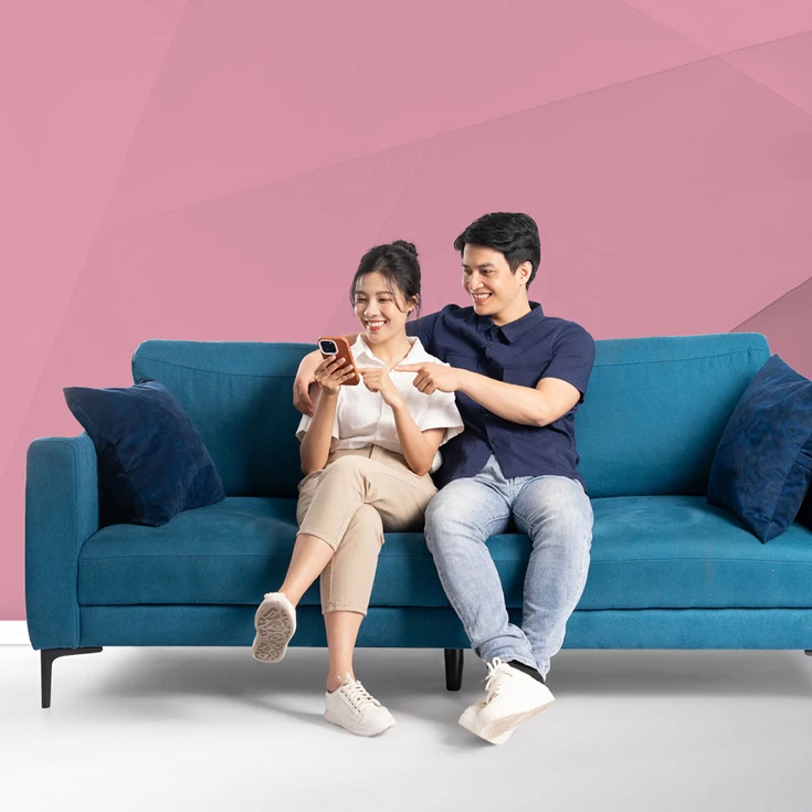 A cheerful couple sitting on a blue sofa, smiling and engaging with a smartphone, symbolising connection and shared experiences, against a blue geometric background.