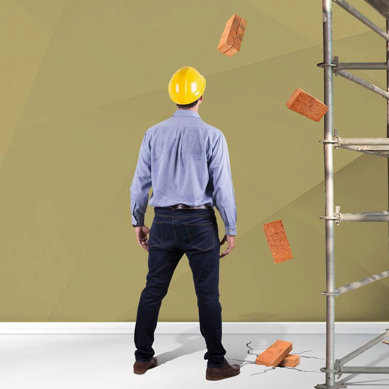 A construction worker wearing a yellow hard hat and a blue shirt stands facing a partially collapsing wall, with bricks falling to the ground. A scaffold is visible to the right. The background features a muted yellow geometric design. Text on the left reads ‘Repair Assist’ followed by ‘When you need experts to put right what’s gone wrong.’