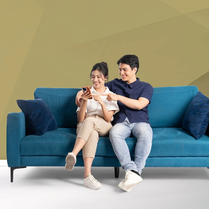 A cheerful couple sitting on a blue sofa, smiling and engaging with a smartphone, symbolising connection and shared experiences, against a blue geometric background.