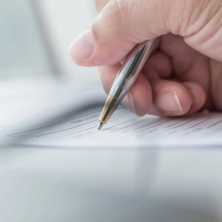 Close-up of a hand holding a pen, writing on a sheet of paper with neatly ruled lines, symbolising documentation, note-taking, or signing an important document.